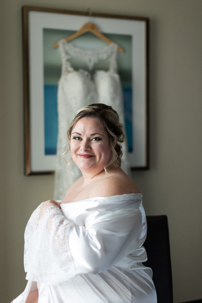 Bride seated with a Pierra's Bridal wedding dress hanging in the background, Lansing Wedding Photographer